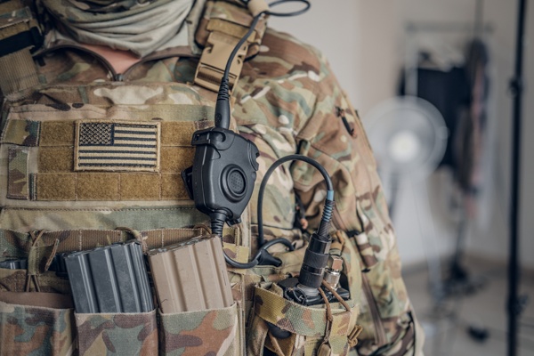 Detail of a soldier's tactical vest featuring an attached communication device with a PTT button and headset connector.
